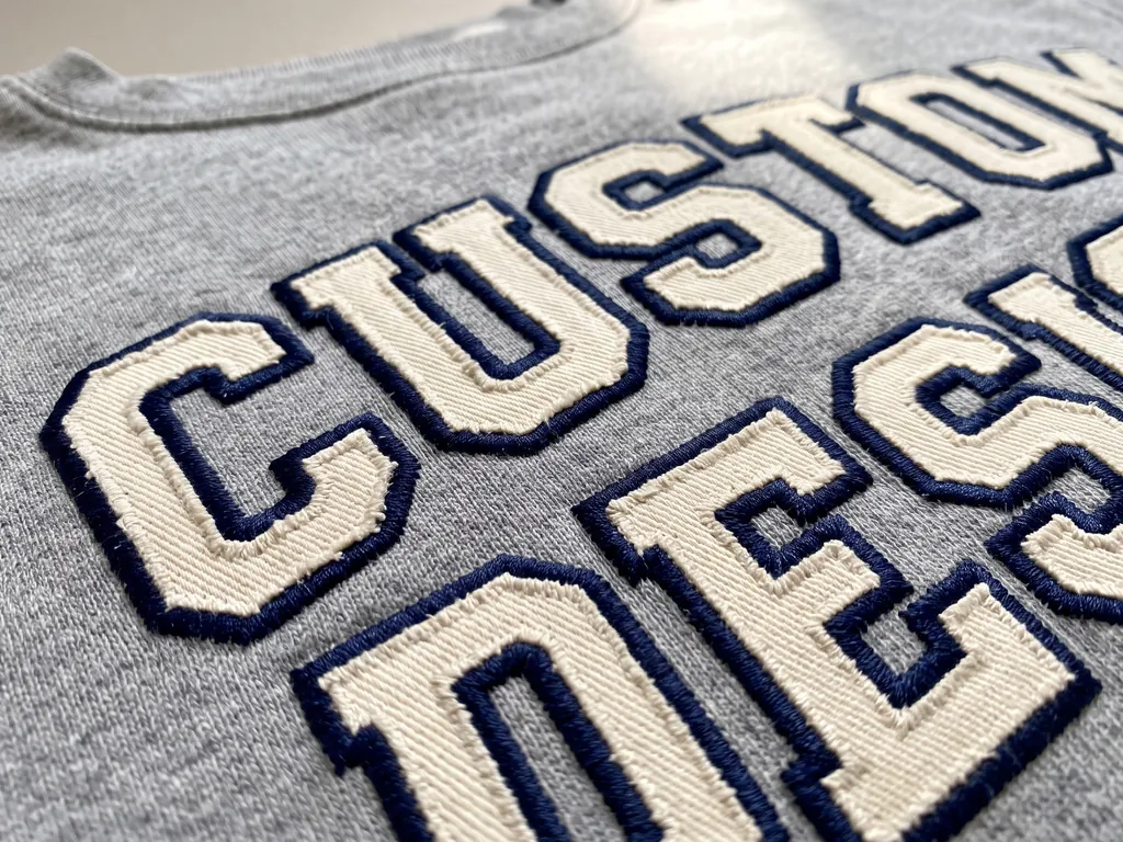 Applique embroidery close-up: cream twill CUSTOM DESIGN letters with navy satin borders on grey marl fleece