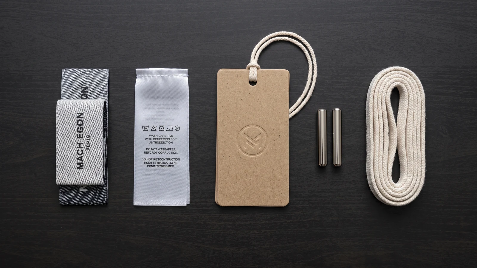 Flat-lay of jogger trims: woven waistband label, satin wash tag, embossed swing tag, metal drawcord tips and a flat drawcord