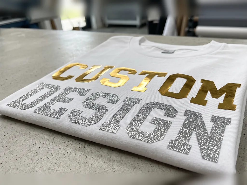 Gold metallic and silver glitter vinyl transfers spelling CUSTOM DESIGN on a folded white t-shirt